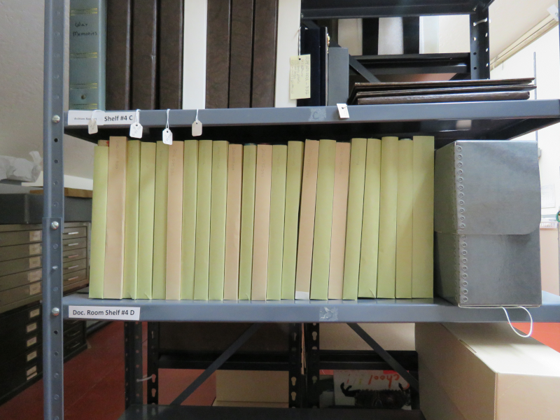 A row of slim books standing upright on a metal shelf, each in a individual cardstock sleeve.